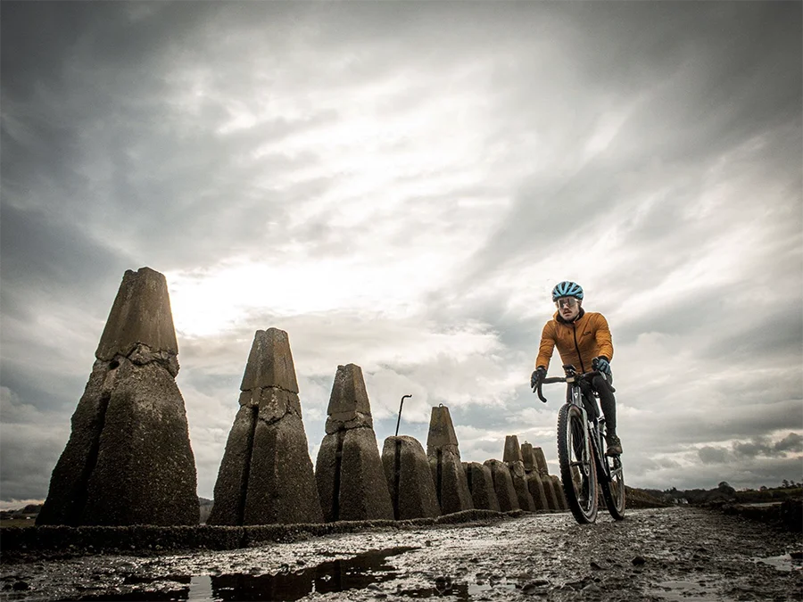 Markus Stitz wearing an orange jacket and blue helmet rides along a wet, muddy path beside a row of large, weathered concrete sea defenses under a cloudy sky.