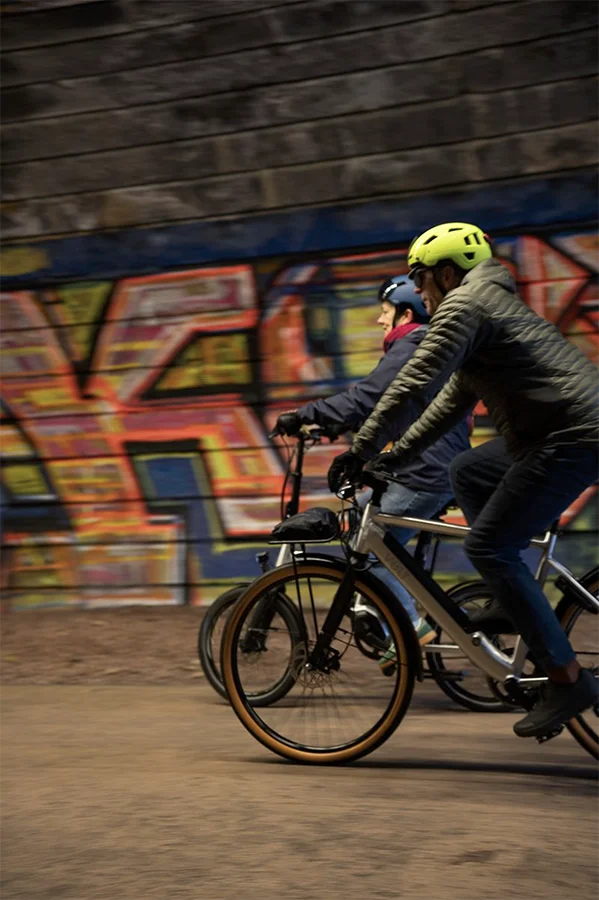 Two cyclists wearing helmets ride side by side past a colorful graffiti-covered wall, with motion blur emphasizing their speed.
