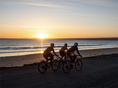 Three cyclists ride along a coastal road at sunset, silhouetted against the ocean and glowing horizon.